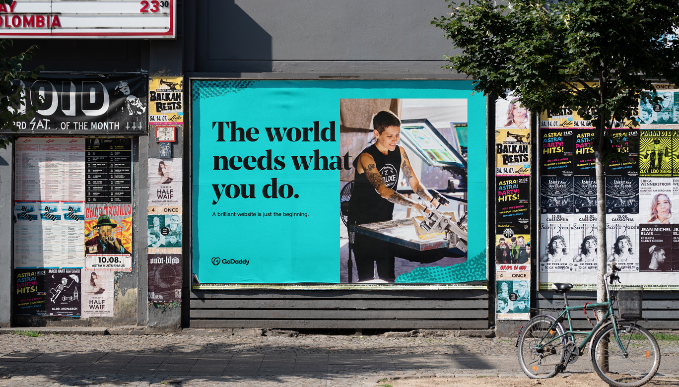 Blue colored billboard of GoDaddy Design showing a small scale entrepreneur working