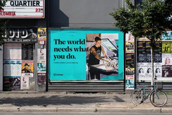 Blue colored billboard of GoDaddy Design showing a small scale entrepreneur working